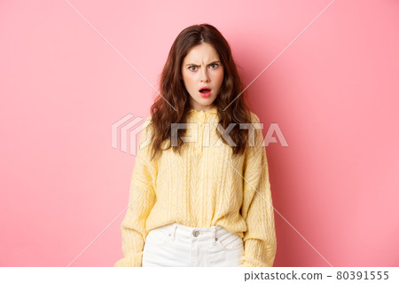 Shocked and confused brunette woman gasping, frowning and looking offended at camera, feeling insulted or displeased, standing over pink background Shocked and confused brunette woman gasping, frowning and looking offended at camera, feeling insulted or displeased, standing over pink background 80391555