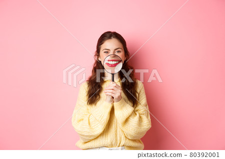 Stomagology and healthcare concept. Young beautiful girl with perfect smile, showing her white teeth with magnifying glass, standing against pink background Stomagology and healthcare concept. Young beautiful girl with perfect smile, showing her white teeth with magnifying glass, standing against pink background 80392001
