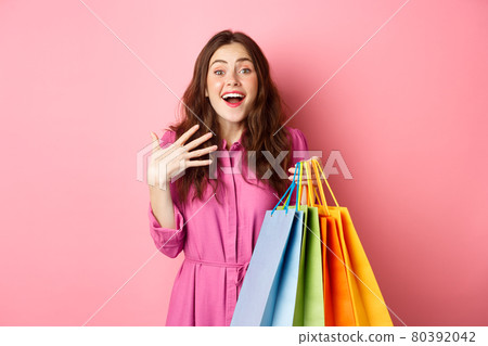 Image of surprised young woman gasping amazed, holding hand on chest and smiling astonished with sale discounts, holding shopping bags, standing against pink background Image of surprised young woman gasping amazed, holding hand on chest and smiling astonished with sale discounts, holding shopping bags, standing against pink background 80392042