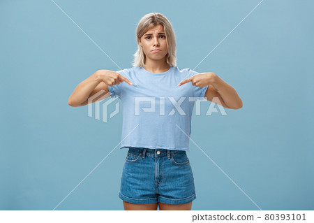 Sad cute blonde young female complaining being displeased with own body, pointing at breast frowning making gloomy smile pulling lips down from disappointment standing over blue background Sad cute blonde young female complaining being displeased with own body, pointing at breast frowning making gloomy smile pulling lips down from disappointment standing over blue background 80393101