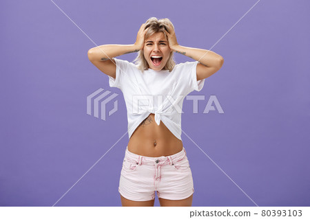 Portrait of panicking concerned and depressed young european woman with blond hair tattoos and athletic body yelling out loud holding hands on head being concerned and shook over purple background Portrait of panicking concerned and depressed young european woman with blond hair tattoos and athletic body yelling out loud holding hands on head being concerned and shook over purple background 80393103