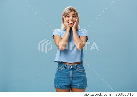 Oh my so cute. Portrait of touched tender good-looking athletic woman with fair hair holding palms on cheeks smiling being impressed and pleased standing in denim shorts over blue background Oh my so cute. Portrait of touched tender good-looking athletic woman with fair hair holding palms on cheeks smiling being impressed and pleased standing in denim shorts over blue background 80393316