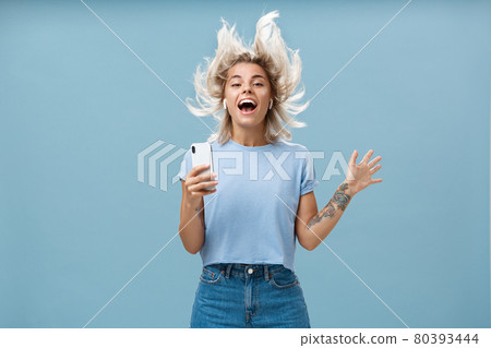 Expressing happiness with help of great tunes. Joyful amused and happy good-looking young female student jumping having fun listenign music in wireless earbuds, holding smartphone over blue wall 80393444