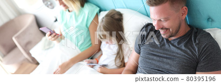 Parents and child lie in bed, each holding smartphone in his hands 80395196