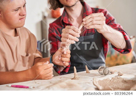 Man with special needs working with clay in ceramic studio under supervision of craftsman Man with special needs working with clay in ceramic studio under supervision of craftsman 80396264