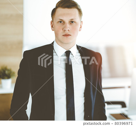 Cheerful businessman standing and looking at camera in sunny office. Headshot of young entrepreneur Cheerful businessman standing and looking at camera in sunny office. Headshot of young entrepreneur 80396971