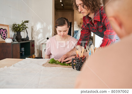 Sculptor showing how to do impression of leaf relief to cute girl with down syndrome 80397002
