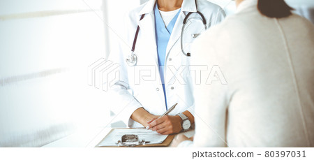 Unknown woman-doctor and patient in sunny hospital. Physician controls medication history records and exam results while using clipboard, close-up Unknown woman-doctor and patient in sunny hospital. Physician controls medication history records and exam results while using clipboard, close-up 80397031
