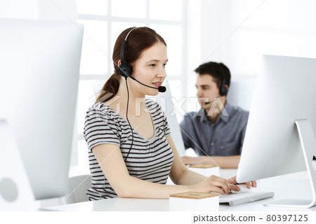 Casual dressed young woman using headset and computer while talking with customers online. Group of operators at work. Call center, business concept 80397125