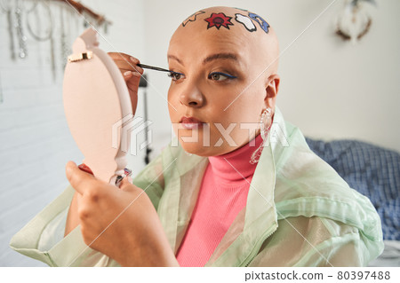 Blogger with patterns at her bald head looking at the hands mirror and preparing make up Blogger with patterns at her bald head looking at the hands mirror and preparing make up 80397488