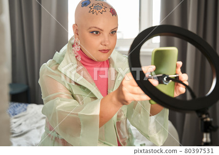 Bald woman sitting in front of the smartphone on tripod and preparing to recording video 80397531