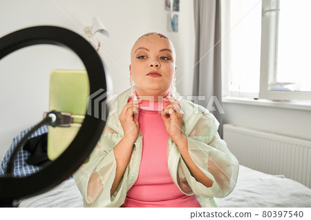 Bald woman sitting on the bed and looking at the camera of a smartphone on tripod Bald woman sitting on the bed and looking at the camera of a smartphone on tripod 80397540