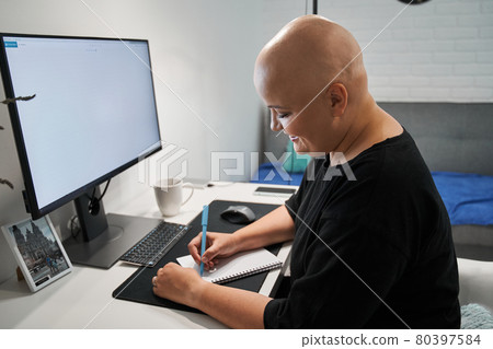 Woman without hair making some notes while doing her work task at the table Woman without hair making some notes while doing her work task at the table 80397584