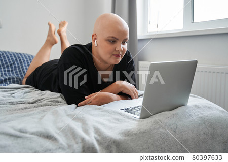 Bald woman wearing earphones working at the laptop while laying on the bed Bald woman wearing earphones working at the laptop while laying on the bed 80397633