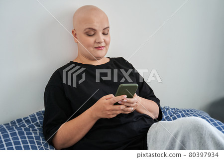 Bald woman laying at the bed and looking at her smartphone while relaxing Bald woman laying at the bed and looking at her smartphone while relaxing 80397934