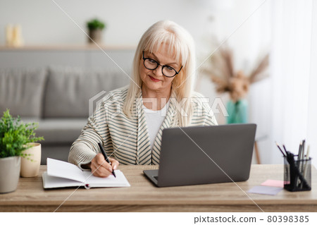 Mature lady writing in notebook and using laptop at home 80398385