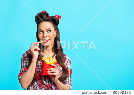 Young pinup woman in retro outfit eating french fries from package, looking aside at empty space on blue background 80398386