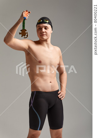 Swimmer with a medal on a gray background rejoices in victory, an athlete in excellent physical shape celebrates a victory clutching a medal in his hand, victory concept, gray background, copy space 80400221