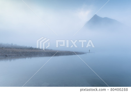 Traveler man standing alone in blue fog with rocky mountains by the lake in the morning 80400288