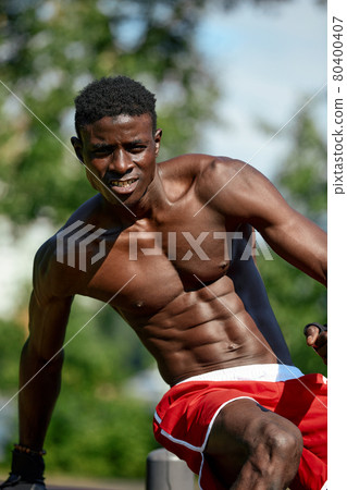 Muscular young man exercising at the sports field. African man looking to the side while doing horizontal bar exercises. Shirtless male model exercising outdoors. Muscular young man exercising at the sports field. African man looking to the side while doing horizontal bar exercises. Shirtless male model exercising outdoors. 80400407