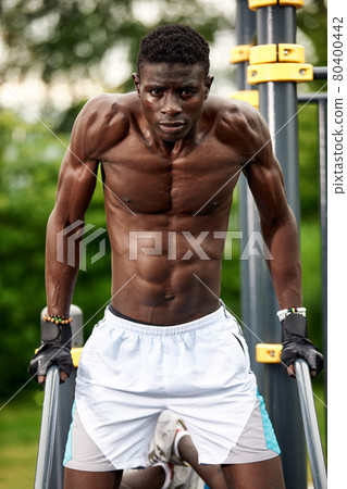 Muscular young man exercising at the sports field. African man looking to the side while doing horizontal bar exercises. Shirtless male model exercising outdoors. 80400442