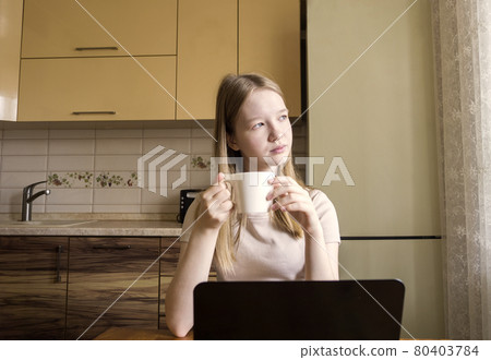 A girl with blonde hair holds a mug of tea in her hands and looks away. The teenager next to the window in front of the laptop is distracted . Education at home, leisure. 80403784
