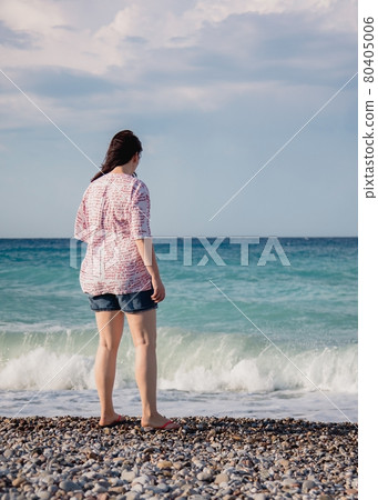 young woman in shorts and a shirt stands on a pebble beach and looks at the waves of the mediterranean sea, rear view, hair fluttering in the wind young woman in shorts and a shirt stands on a pebble beach and looks at the waves of the mediterranean sea, rear view, hair fluttering in the wind 80405006