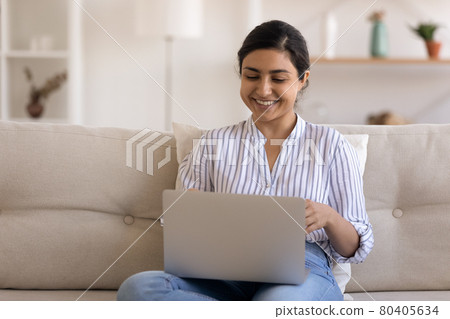 Smiling Indian woman using laptop, having fun, sitting on couch Smiling Indian woman using laptop, having fun, sitting on couch 80405634