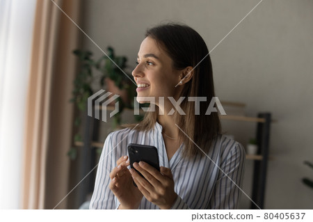 Dreamy beautiful woman holding smartphone standing indoor looks out window Dreamy beautiful woman holding smartphone standing indoor looks out window 80405637