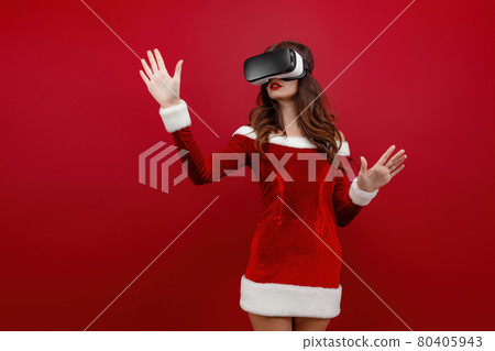 Amazed young Santa girl in Christmas dress looking in headset pointing hands aside isolated on red wall background.  80405943