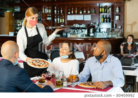 Polite waitress in protective mask bringing ordered pizza to friends visited restaurant 80409992