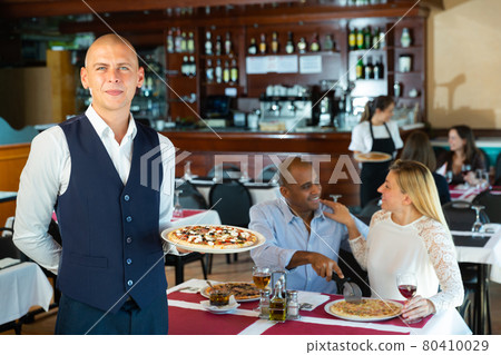 Portrait of smiling waiter in uniform with serving tray pizza at restaurant Portrait of smiling waiter in uniform with serving tray pizza at restaurant 80410029