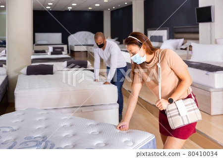 Young Hispanic woman in medical face mask looking for new mattress for bed in furniture store. Young Hispanic woman in medical face mask looking for new mattress for bed in furniture store. 80410034