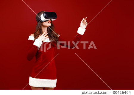 Amazed young Santa girl in Christmas dress looking in headset pointing hands aside isolated on red wall background.  80416464