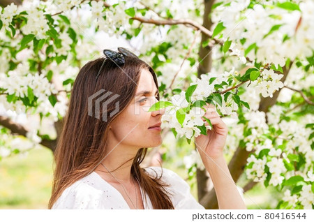 Smiling and happy female is posing in white clothes. Cheerful pretty woman on apple tree background. Young woman caucasian ethnicity. Attractive and beautiful model. Blooming apple trees background Smiling and happy female is posing in white clothes. Cheerful pretty woman on apple tree background. Young woman caucasian ethnicity. Attractive and beautiful model. Blooming apple trees background 80416544