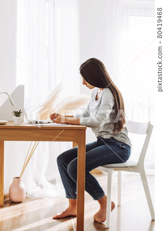 A beautiful student girl is sitting at a desk and making notes in a notebook, using a laptop to prepare for classes. Modern technologies, online education, training 80419468