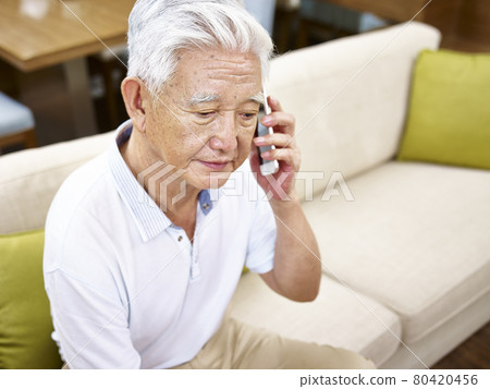 senior asian man sitting alone on sofa talking on mobile phone looking sad 80420456