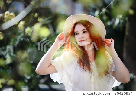 Portrait of young woman with hat standing outdoors in city in summer. 80420954