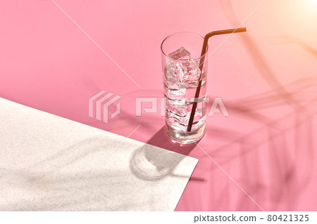 Glass of mineral water with ice on pink and white background with shadow from a palm leaf. Sun flare Glass of mineral water with ice on pink and white background with shadow from a palm leaf. Sun flare 80421325