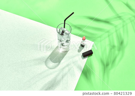 High angle view of lipstick and drinking glass with ice on table. Green and white background with shadow from a palm leaf 80421329