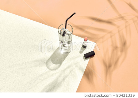 High angle view of lipstick and drinking glass with ice on table. Beige and white background with shadow from a palm leaf 80421339
