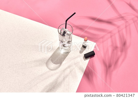 High angle view of lipstick and drinking glass with ice on table. Pink and white background with shadow from a palm leaf 80421347