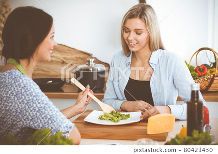Two women friends taste new recipes for delicious fresh salads while sitting at the kitchen table. Vegetarian cooking concept 80421430