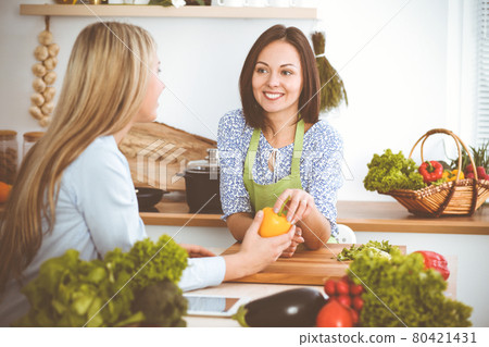 Two women friends choose the recipe and ingredients for a delicious meal while sitting at the kitchen table. Vegetarian concept 80421431