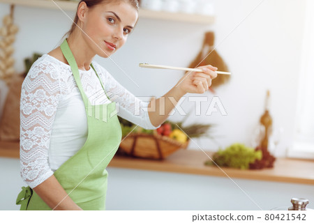 An attractive young dark-haired woman frying meal at worm pan by new keto recipe while standing and smiling in sunny kitchen. Cooking and householding concepts 80421542