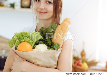 An attractive young woman holding the paper bag full of vegetables while standing and smiling in sunny kitchen. Cooking concept An attractive young woman holding the paper bag full of vegetables while standing and smiling in sunny kitchen. Cooking concept 80421545