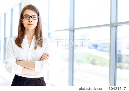 Beautiful female specialist with arms crossed standing in modern office and smiling charmingly. Working on design, data analysis, plan strategy. Business people concept Beautiful female specialist with arms crossed standing in modern office and smiling charmingly. Working on design, data analysis, plan strategy. Business people concept 80421547