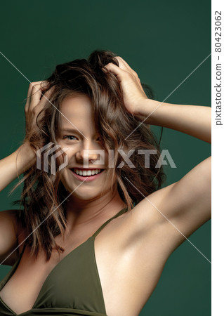 Portrait of young beautiful tanned smiling woman in green lingerie, swimming suit posing isolated over dark green studio background. Natural beauty 80423062