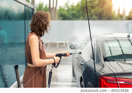 Side view young adult woman washing car with high pressure water equipment pump self-service outdoor on bright windy summer day. Vehicle covered foam shampoo chemical detergents carwash self service 80423686