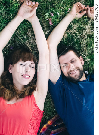 Couple makes a selfie while lying on the grass in flowers. Rest, lovers on a flower meadow. Bearded man and a girl in a red dress take pictures of themselves in the field. Husband and wife on vacation 80423709